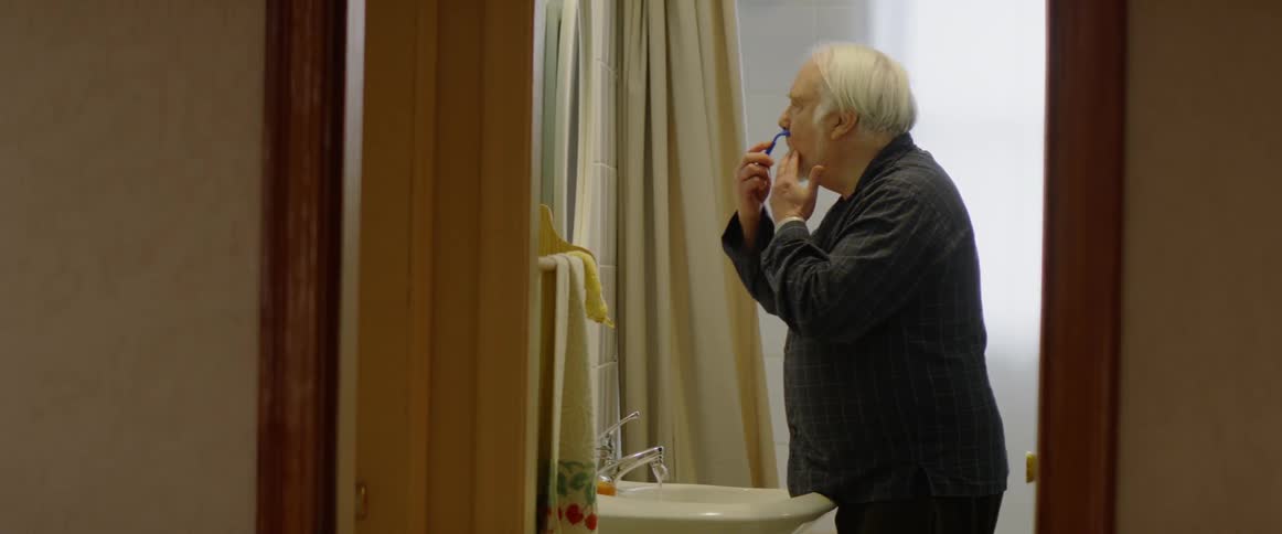 An elderly man shaves in a bathroom mirror