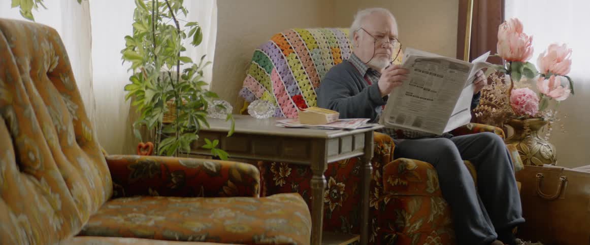 Elderly man wearing glasses sits in a chair reading a newspaper