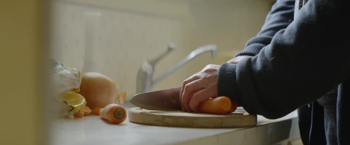 An old man is chopping carrots up on a board in a kitchen in daylight
