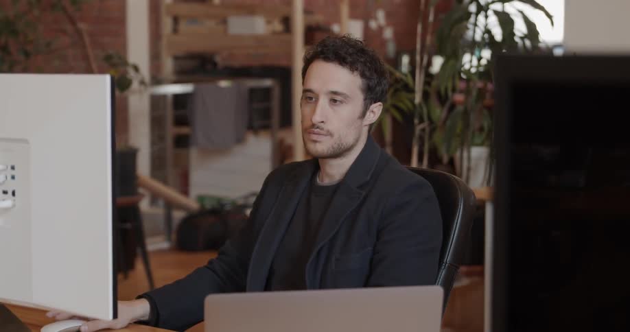 A man sits at a desk with a laptop and computer monitor