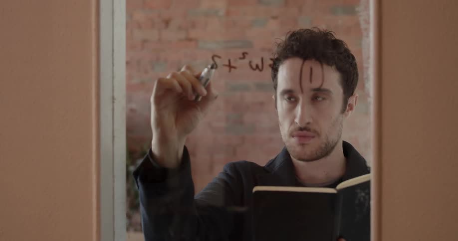 A man is writing out an equation on a glass board inside in daylight