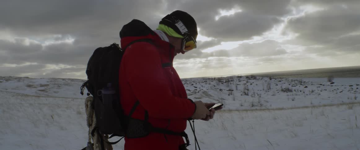 A man is pulling out and using his cellphone while trekking in a remote area on a winter day