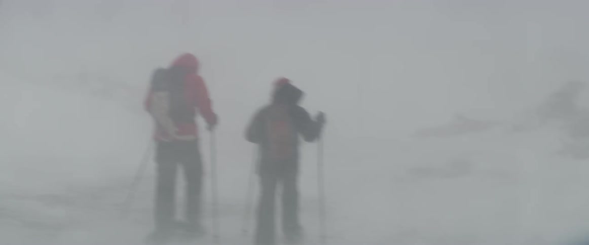 Two people are trekking through a blizzard in a remote area in daylight