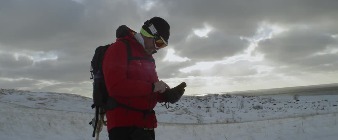 Man wearing goggles and a backpack stands in the snow and looks at a mobile device