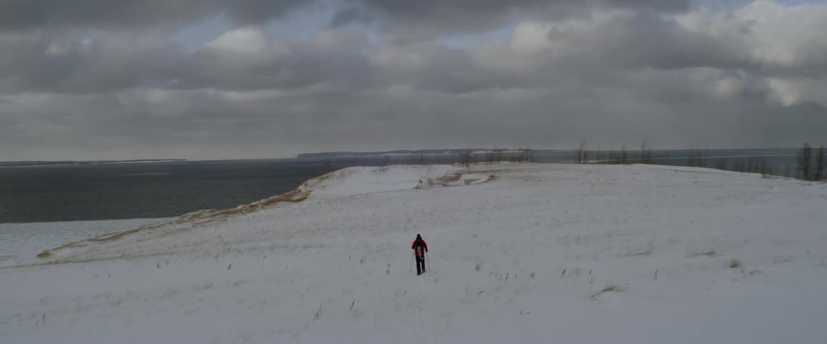 A man is trekking across a snow-covered landscape toward the coast in daylight