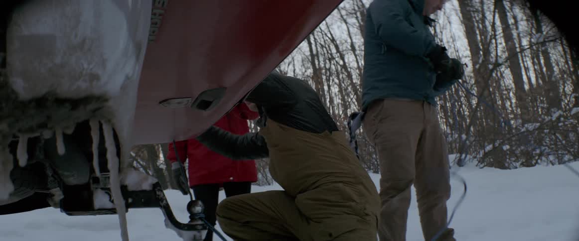 A man is tying a rope to the underside of a vehicle as he works with others outside in winter