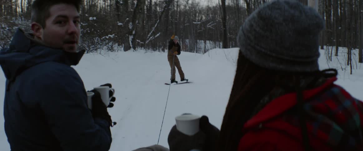And and woman holding mugs look out at a guy holding onto a rope snowboarding