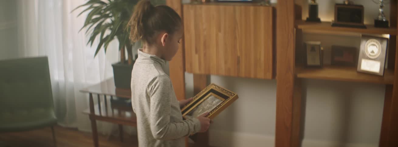 A young girl standing in a living room holding and looking at an old photograph of a woman