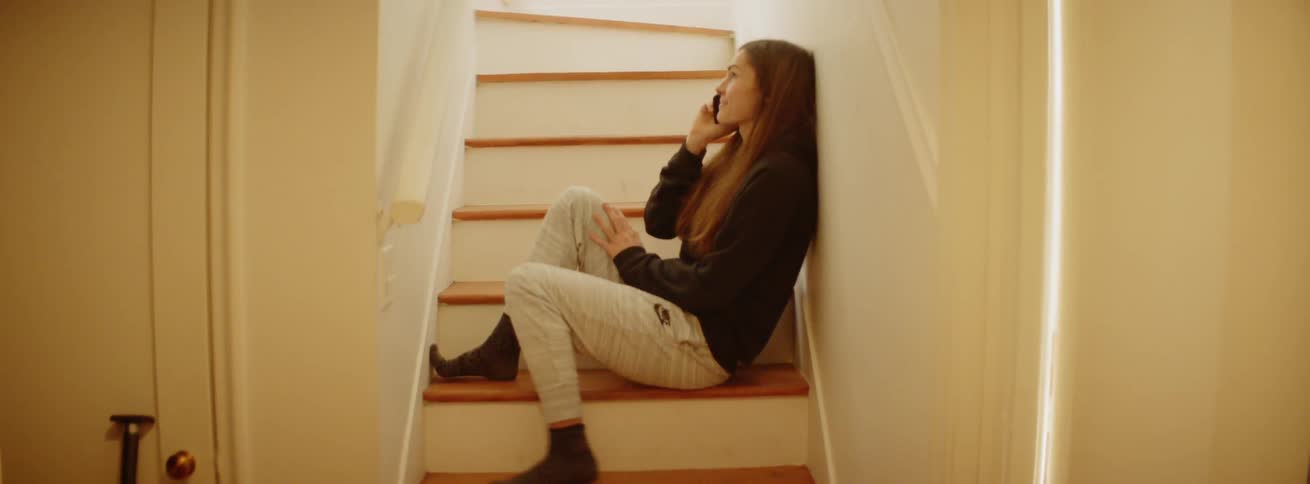 A woman is sitting on a staircase in her house in daylight speaking on a phone
