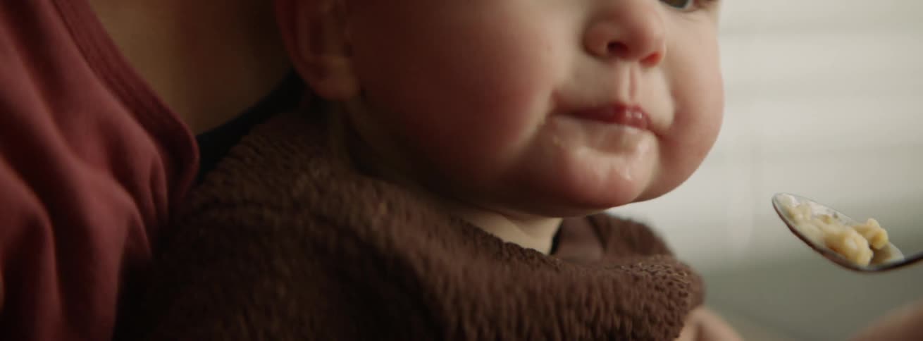 A woman holds a baby in her lap and spoon feeds it