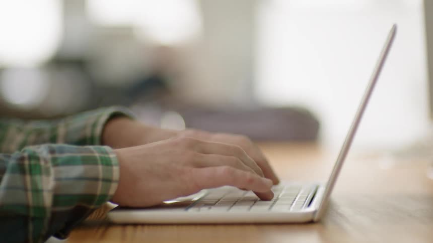 A man is typing on a laptop inside during daylight