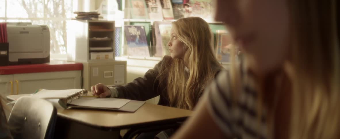 A young, blonde girl sitting at a school desk, looking out the window and then writing in her notebook