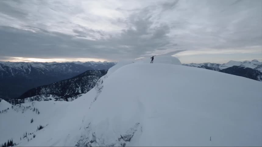 A skier is standing on top of a snow-covered mountain ridge enjoying the view at dusk