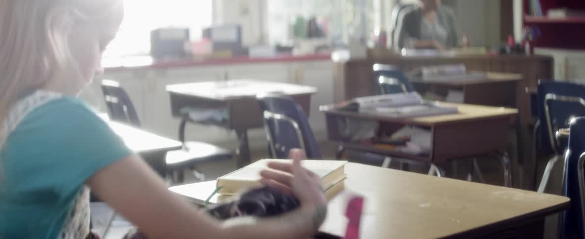 A teacher sits at her desk as a student sits down and starts unpacking her backpack onto her own desk