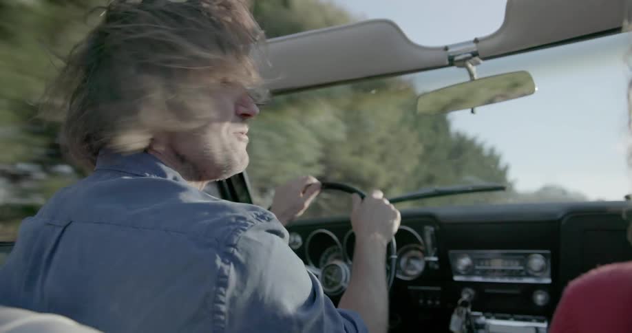 A man's hair is being blown about as he drives in a convertible on a sunny day