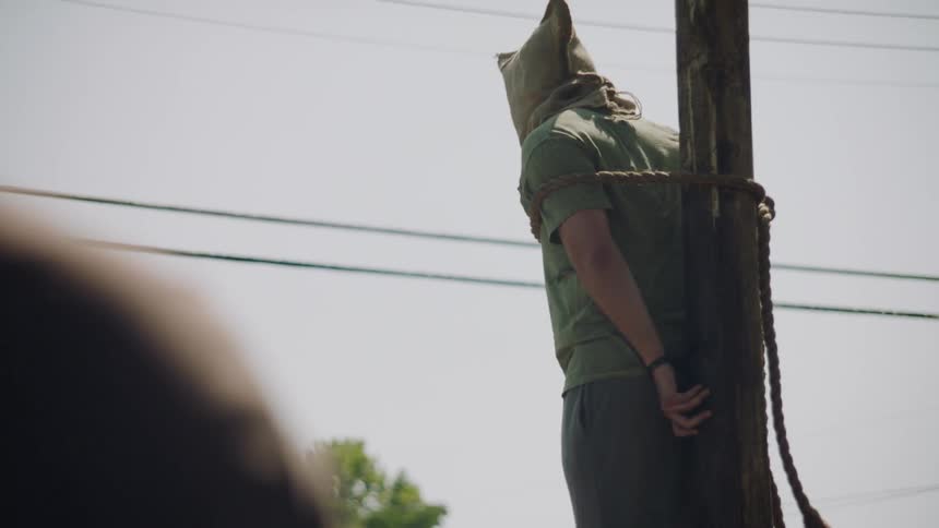 A prisoner with a sack over his head is executed outside in daylight