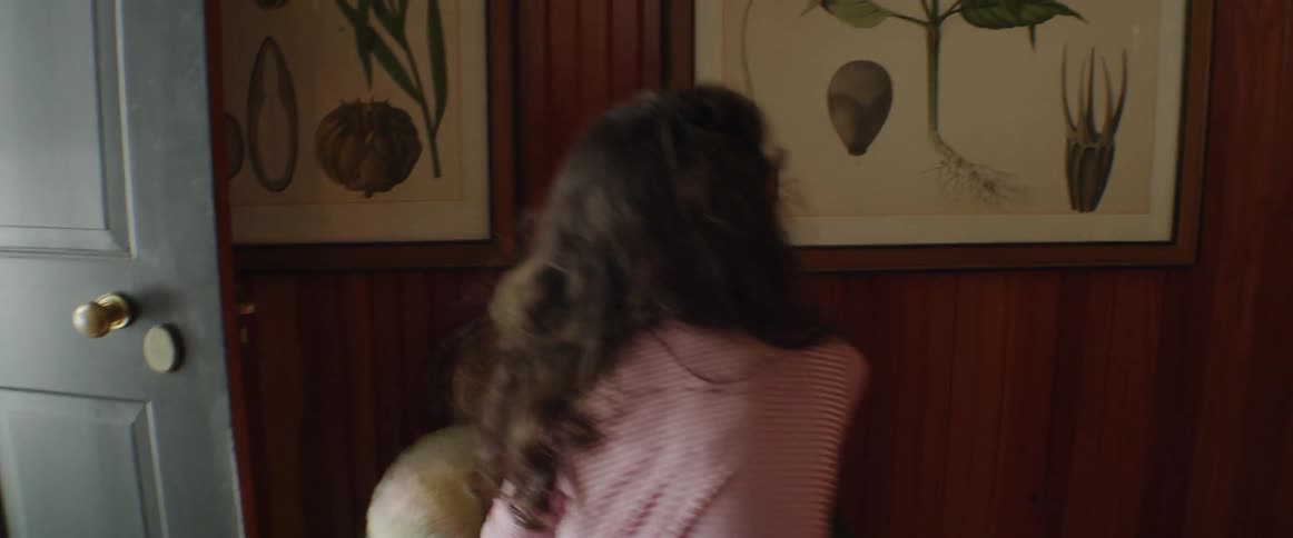 A girl carries a large teddy bear down the stairs and puts it in a chair
