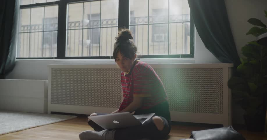 A woman sits on the floor while eating and using a laptop