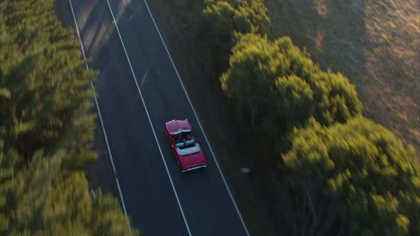 A couple are driving in a convertible along a country road in daylight