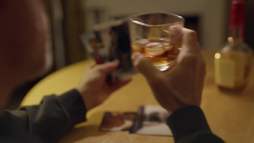 The man is examining photographs at the table before taking a drink