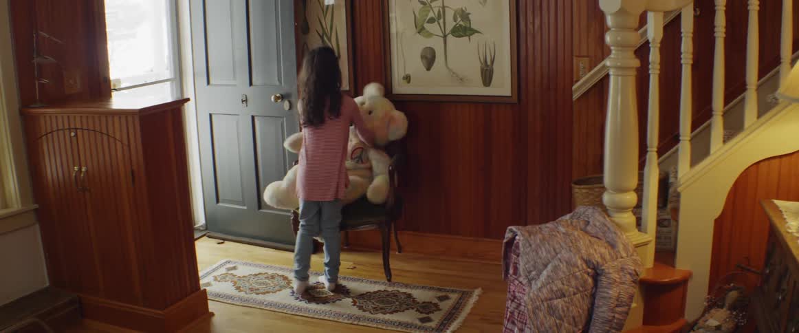 A girl carries a large teddy bear down stairs