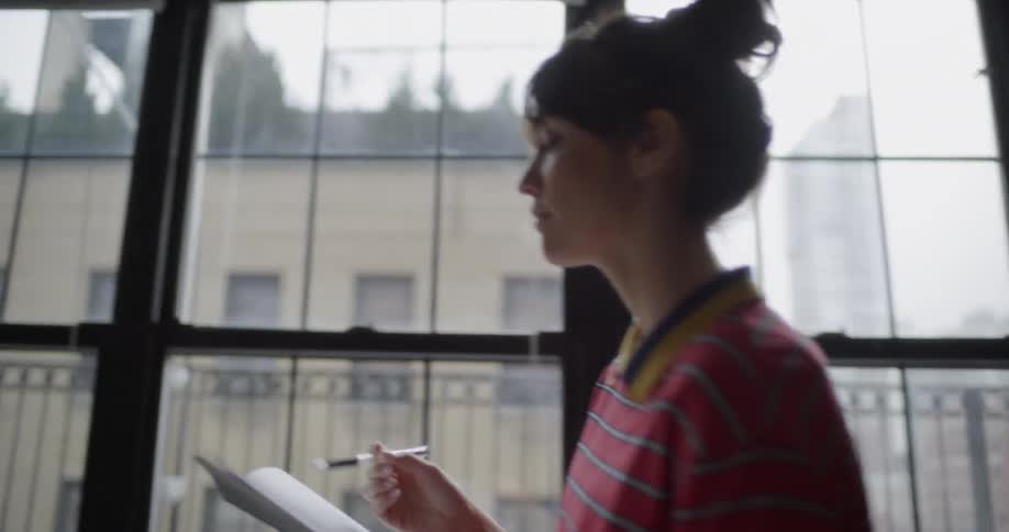 Woman walks in a room with large windows holding a notebook