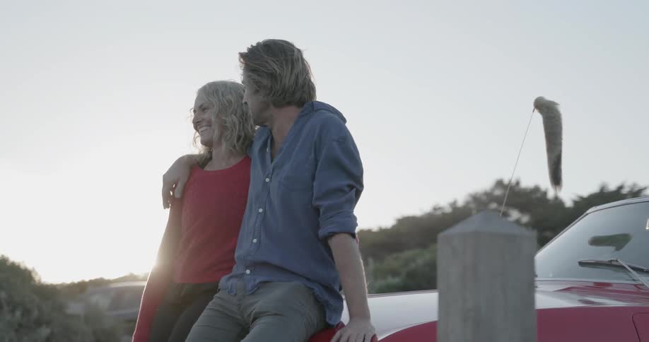 A man and woman smile as they lean on the car together