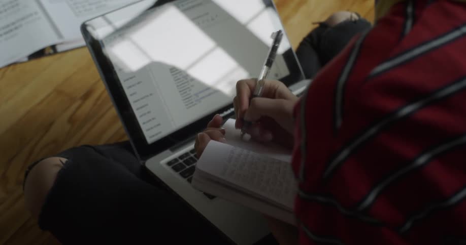 A woman is taking notes in a notebook as she studies on the floor in daylight
