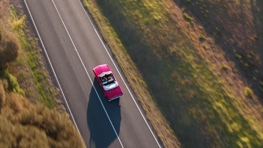 A couple are driving in a convertible along a country road in daylight