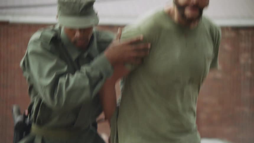 A soldier is leading a prisoner to a pole in a town in daylight