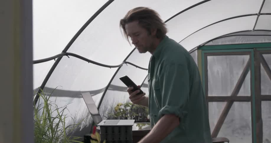 A man in a greenhouse picks up potted plants and photographs them with his phone