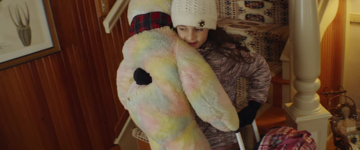 A girl carries a large teddy bear and suitcase outside