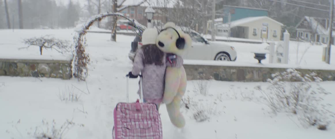 A girl is carrying a stuffed bear and towing a suitcase towards a car on a winter day