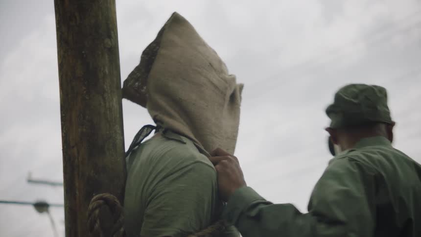 A soldier is putting a sack over a prisoner's head outside on a cloudy day