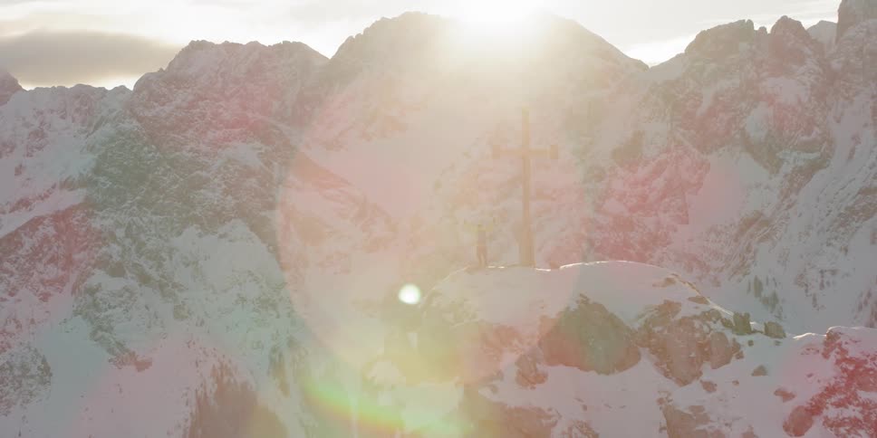 Man standing next to large cross statue on snowy mountain peak