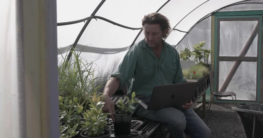The man is sitting on the table in the greenhouse with a laptop