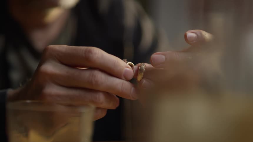 A man holds two wedding rings while drinking whiskey