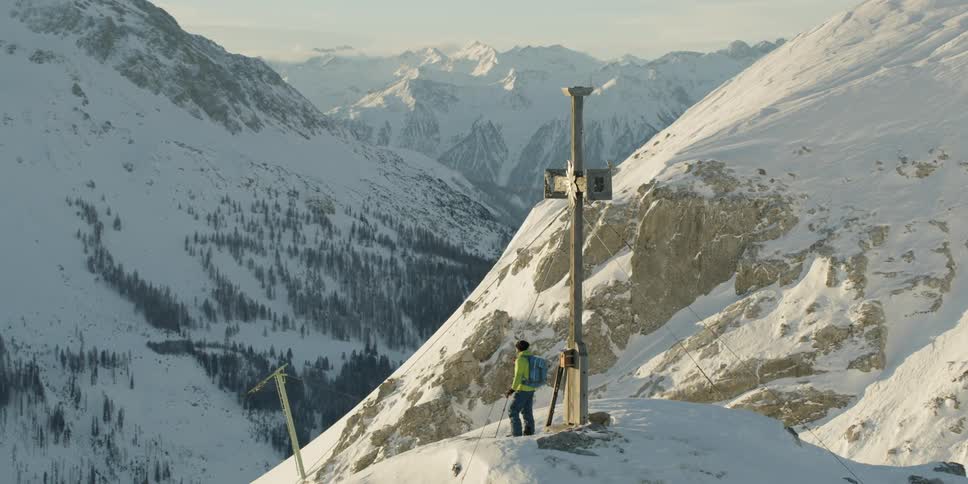 Man stands next to a large cross on a mountain top under a blue sky