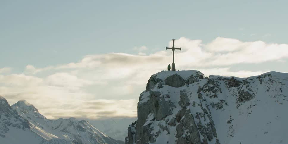 Man and a large cross sit on a snowy mountain under a blue sky