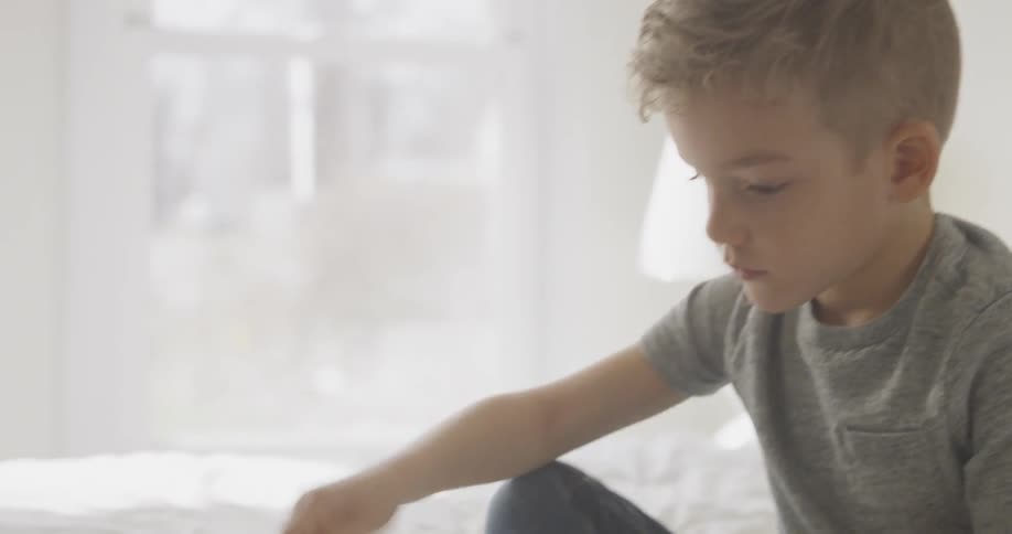 A boy is sitting on his bed playing with lego-style bricks during the day