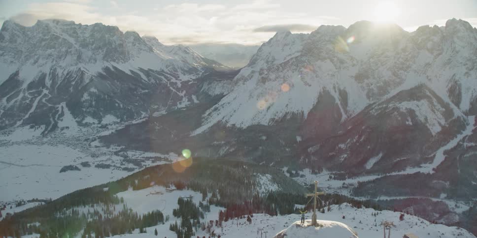A person stands on a snow covered peak next to a cross with arms raised