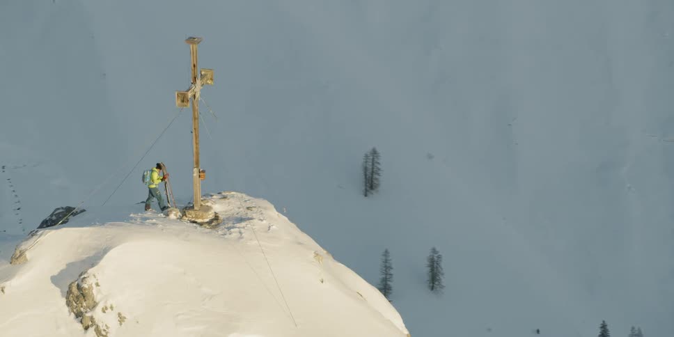 Man with skis stands next to cross statue on top of snow covered mountain peak