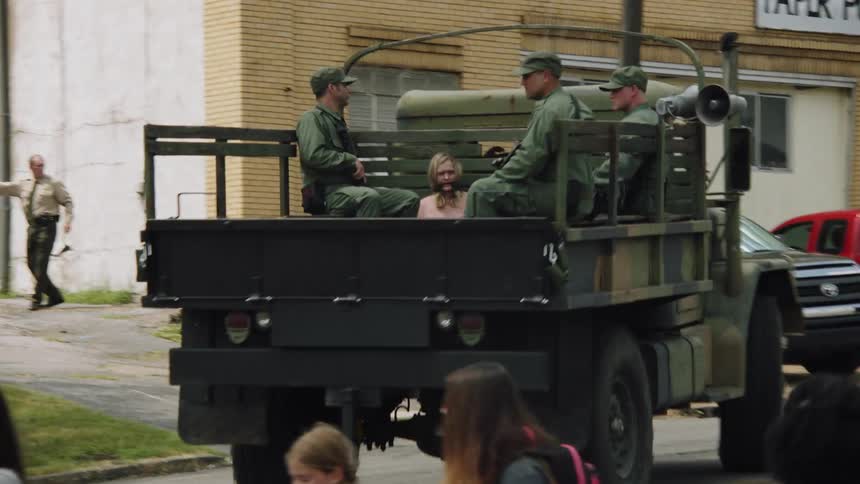 A military truck carrying prisoners is directed down a street during the day