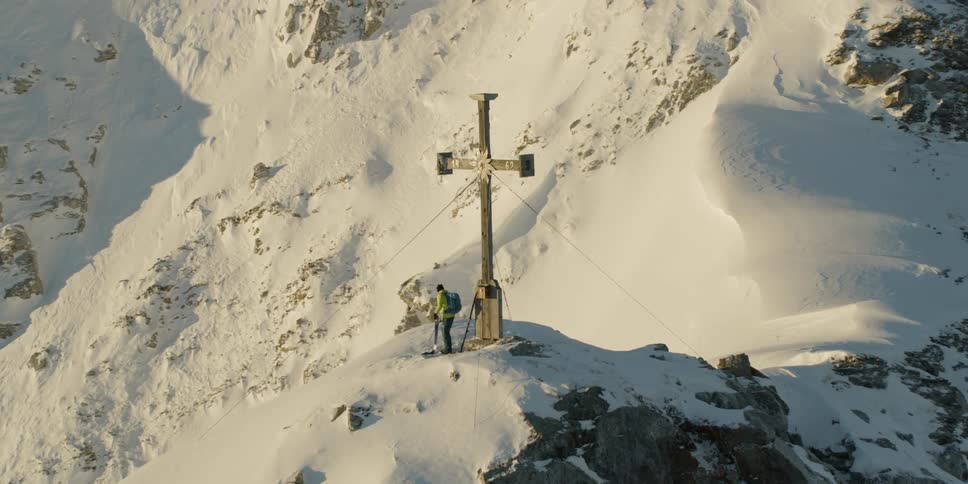 A person with a ski stands by a cross on the mountain