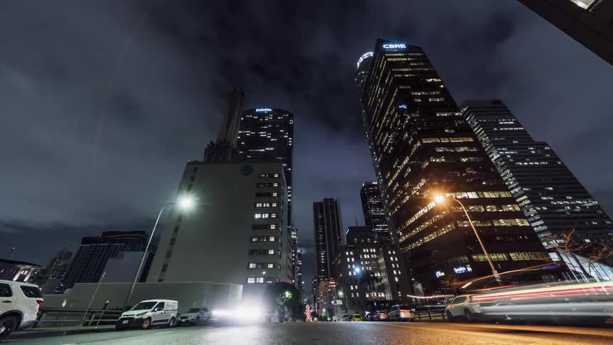 The cars drive past the brightly lit tall buildings