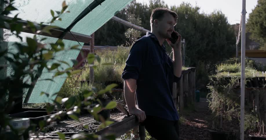 A man leans against a greenhouse table and talks on the phone