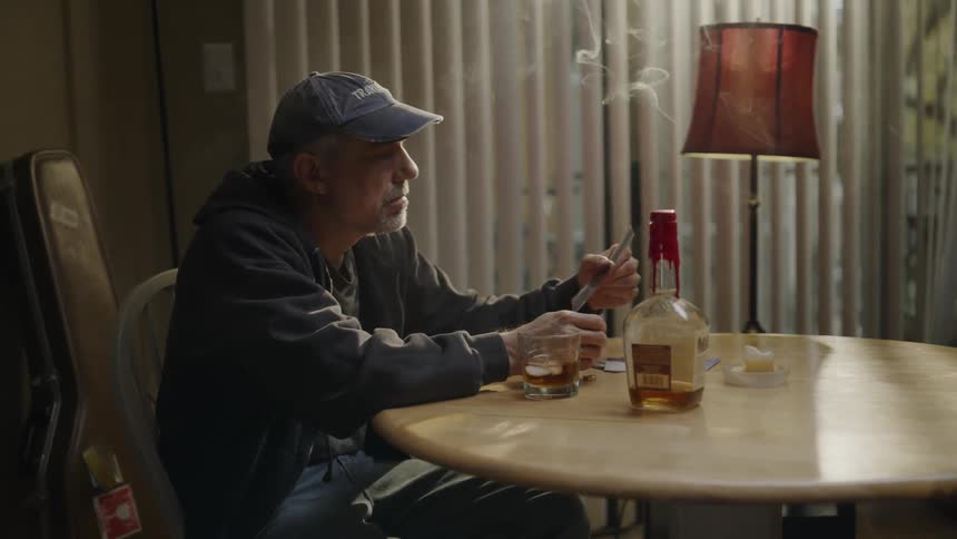 Man sits at table drinking whiskey and smoking a cigarette