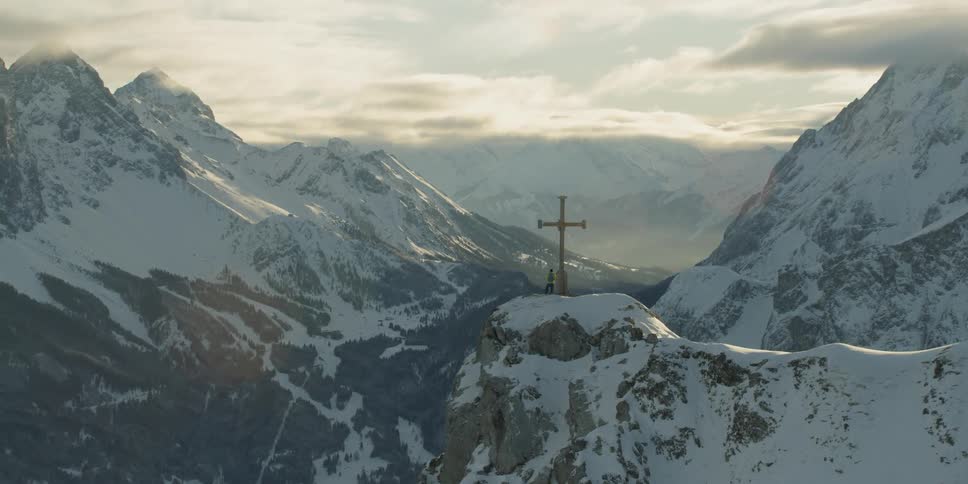 A hiker stands next to a cross on a snowy mountain peak