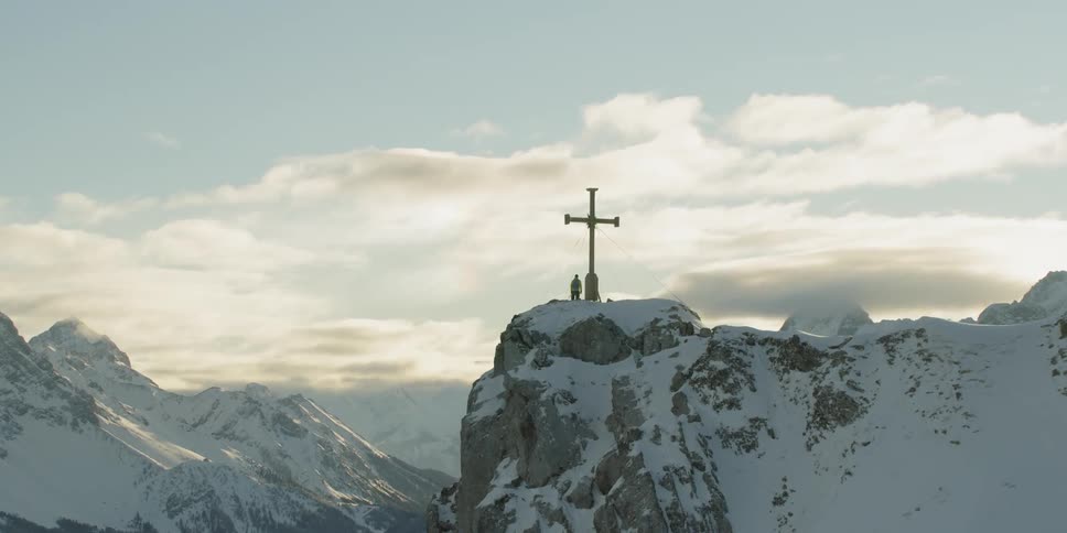 Person stands next to giant cross statue on top of snowy mountain