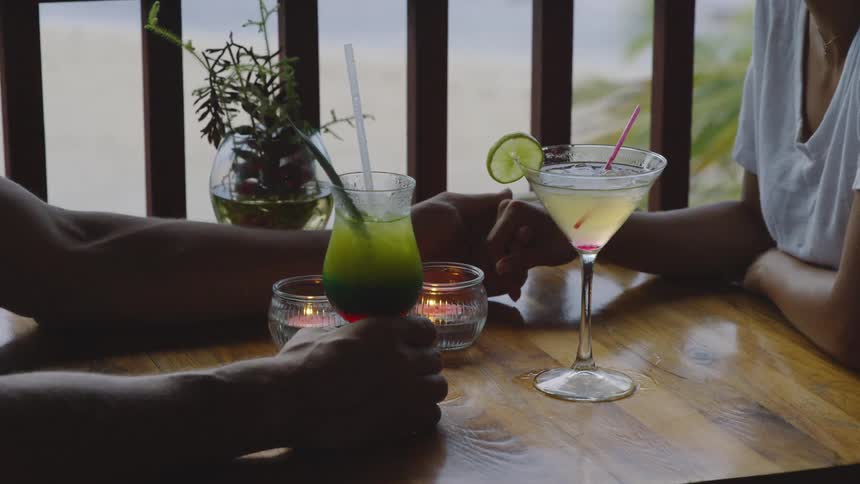 A couple holds hands across the table with tropical drinks on it